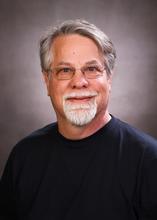 headshot of Evan K. Wallace, a man with short gray hair, a gray beard, and glasses.