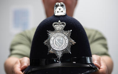 A blurry figure in the background holds out a black police helmet with a silver seal that reads "Norfolk Constabulary."