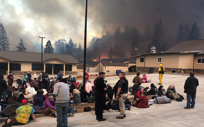 Groups of people sit or lie in a parking lot surrounded by low buildings as fires burn in the forest beyond.  