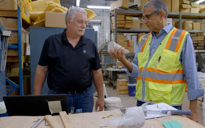 Two men stand a table in a warehouse space, looking at a broken piece of concrete that one is holding. 