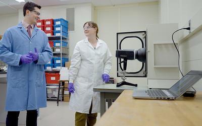 Ryan Falkenstein-Smith and Amy Mensch, both wearing lab coats and gloves, stand in the lab next to a black camera-like device on a table. 