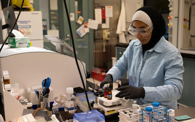 SURF student Zainab Altamimi, who wears a hijab, stands in a lab with safety glasses and gloves on as she holds a micropipette in one hand and a vial in the other.