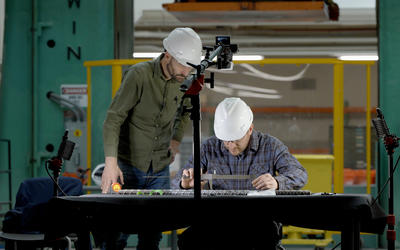 Two researchers in hard hats lean over a rebar sample on a table in a large workspace. 