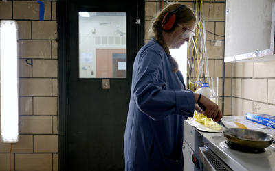 Side view of a woman wearing safety glasses, ear protection, and a blue lab coat. With one hand, she is holding a stainless steel frying pan over an electric stove. With her other hand, she is using a spatula to prod bacon in the pan. The room is smoky.