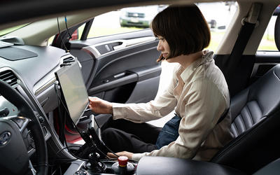 A woman sits in the passenger side of a parked car, looking at a large monitor attached to the dashboard. 