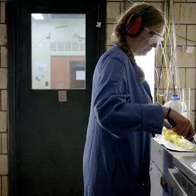 Side view of a woman wearing safety glasses, ear protection, and a blue lab coat. With one hand, she is holding a stainless steel frying pan over an electric stove. With her other hand, she is using a spatula to prod bacon in the pan. The room is smoky.