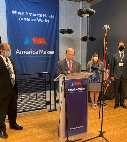 A man stands at a podium labeled "America Makes," with a man and woman at his left and a man on his right.  