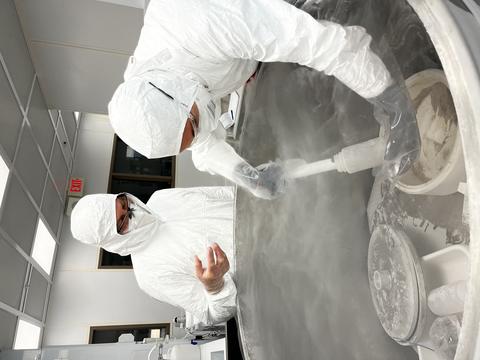 Researchers in white hazmat coveralls reach into an open freezer with a scooping device as vapor rises from the freezer.