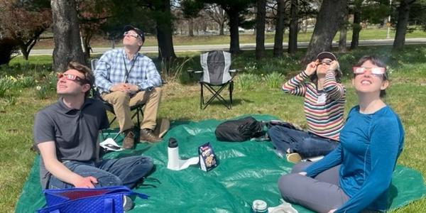 Four people sitting outside on a green tarp wear eclipse glasses as they peer up into the sky. 