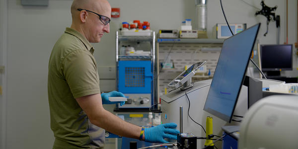 Thomas Forbes wears safety glasses as he looks at the screen attached to a piece of equipment in the lab. 