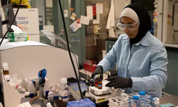 SURF student Zainab Altamimi, who wears a hijab, stands in a lab with safety glasses and gloves on as she holds a micropipette in one hand and a vial in the other.