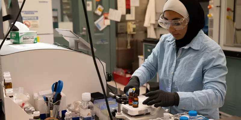 SURF student Zainab Altamimi, who wears a hijab, stands in a lab with safety glasses and gloves on as she holds a micropipette in one hand and a vial in the other.