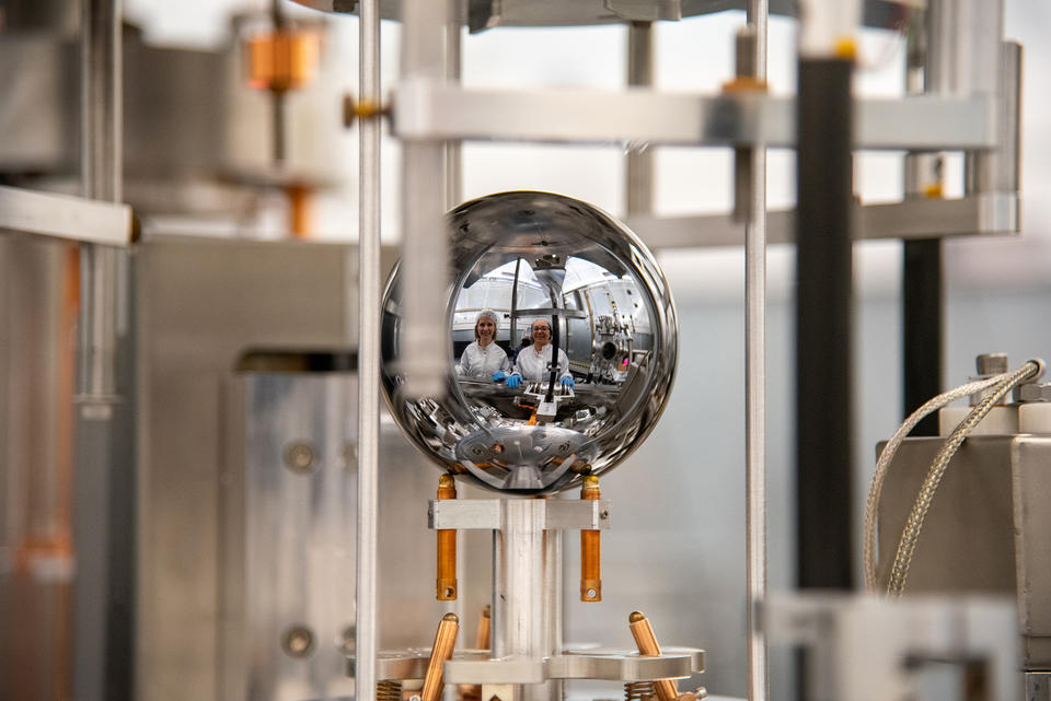 The faces of two researchers in lab coats are reflected in a silvery sphere that is being held inside a larger scientific device. 