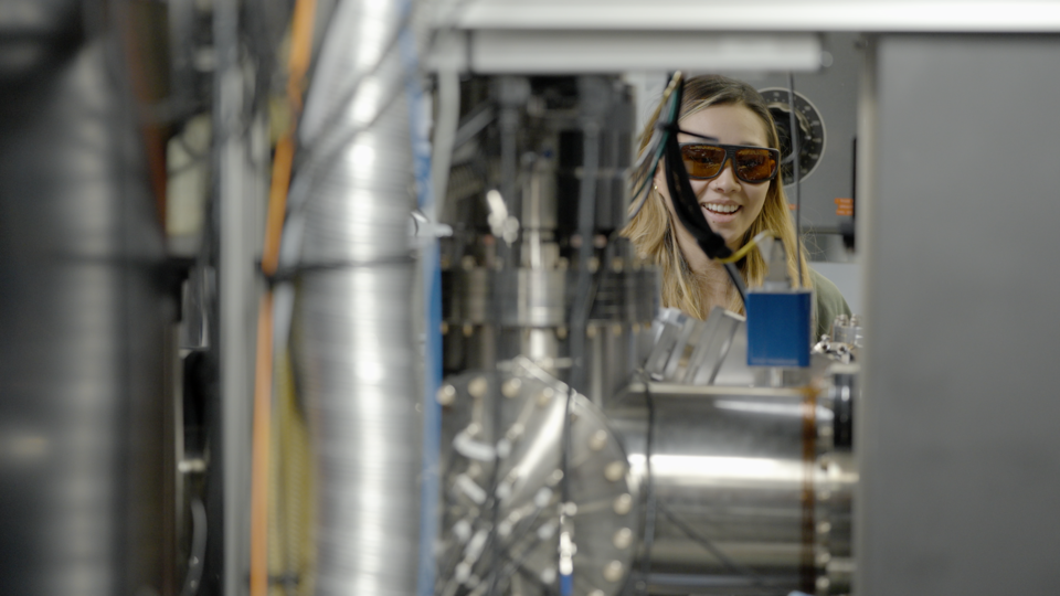 Sun Park, wearing safety glasses, is seen through an opening in an equipment setup in the lab. 