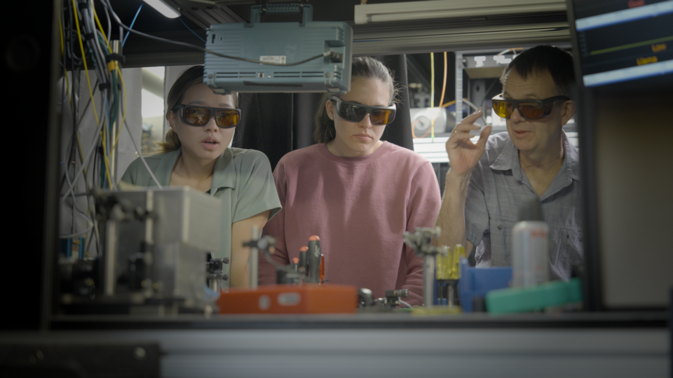Three researchers wearing safety glasses stand in a row looking down at a complex setup of lab equipment. 