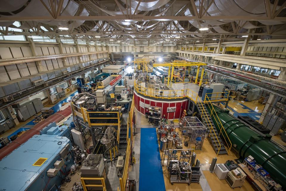 View from near ceiling shows NCNR guide hall filled with big pieces of scientific equipment, including several long cylindrical "guides" running toward the far end of the room. 