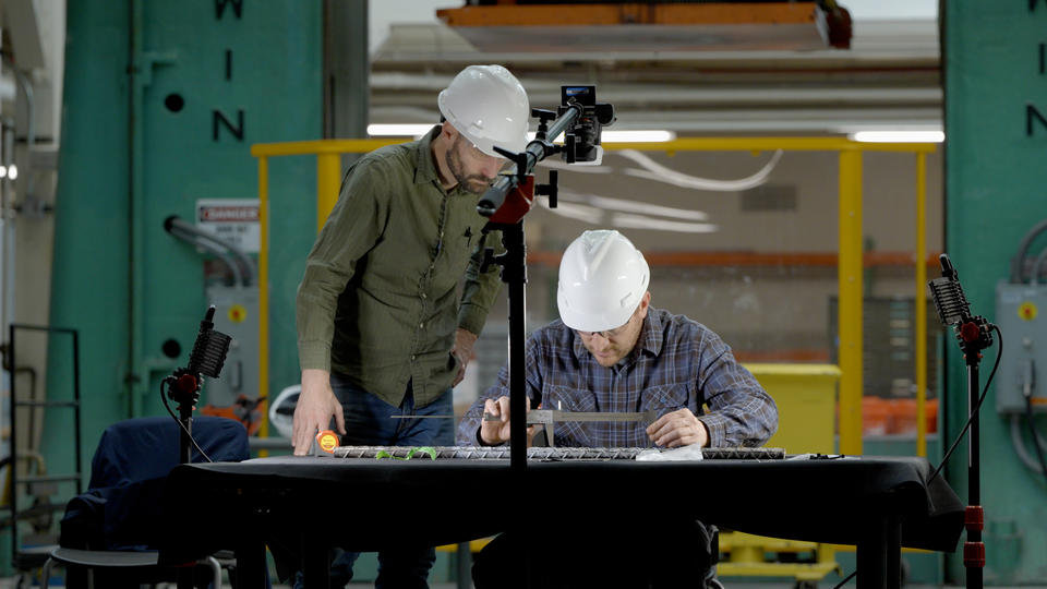 Two researchers in hard hats lean over a rebar sample on a table in a large workspace. 