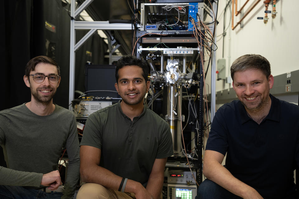 Three male researchers pose smiling in front of a large, complex scientific device in a lab.