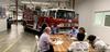 Image of a fire truck inside a firehouse with a group of plain clothes people gathered at a table in discussion