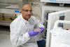 A researcher wearing safety gear kneels at the open door of a lab fridge, displaying a tray of samples. 