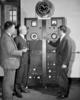 Historical photo shows three men standing next to a refrigerator-sized piece of machinery with an analog clock attached to the top. 