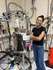 Cassie Stoffer smiles while wearing safety glasses and standing next to a large metal tank with many pipes and wires attached to it. 