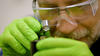 A researcher wearing green gloves and safety glasses over his regular glasses holds a small magnifying glass close to his face to look at a hard drive.