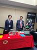 Two men in suits stand behind a display table displaying a laptop and a low cost materials science robot.